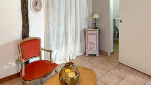 A room with a red upholstered chair, a small table with a decorative vase, a white curtain, and a small cabinet with a lamp. - Beautiful apartment in Puy-L'Evêque (Puy-L'Evêque)