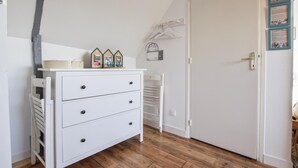 En vit byrå med lådor och en hylla, en vit stol, ett trägolv och en dörr med en ljusströmbrytare och en klädhängare.