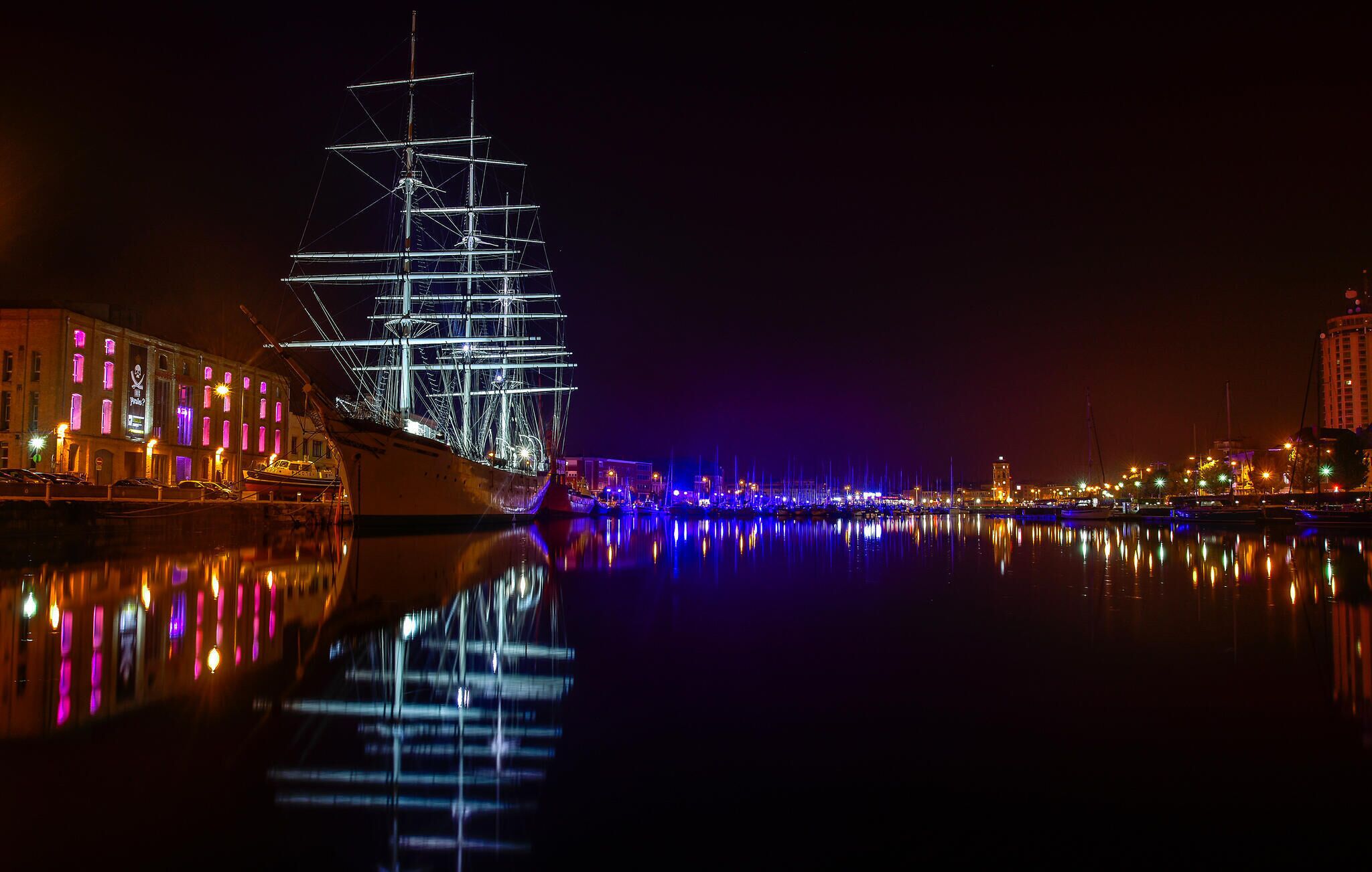 Een groot, verlicht schip afgemeerd in een haven, met gebouwen aan de linkerkant en een stadsgezicht op de achtergrond.