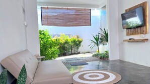 A modern living room with a sofa, a TV mounted on the wall, and a view of a garden. - Altura Esthetic Villas (Batam)
