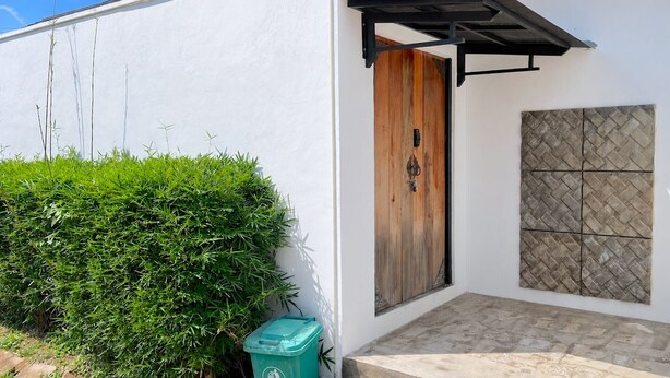 A white building with a wooden door, a green trash bin, and a tiled wall. - Altura Esthetic Villas (Batam)
