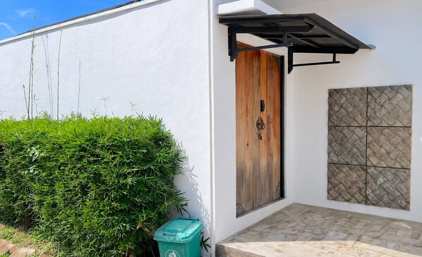 Un edificio blanco con puerta de madera, un cubo de basura verde y un muro con azulejos.