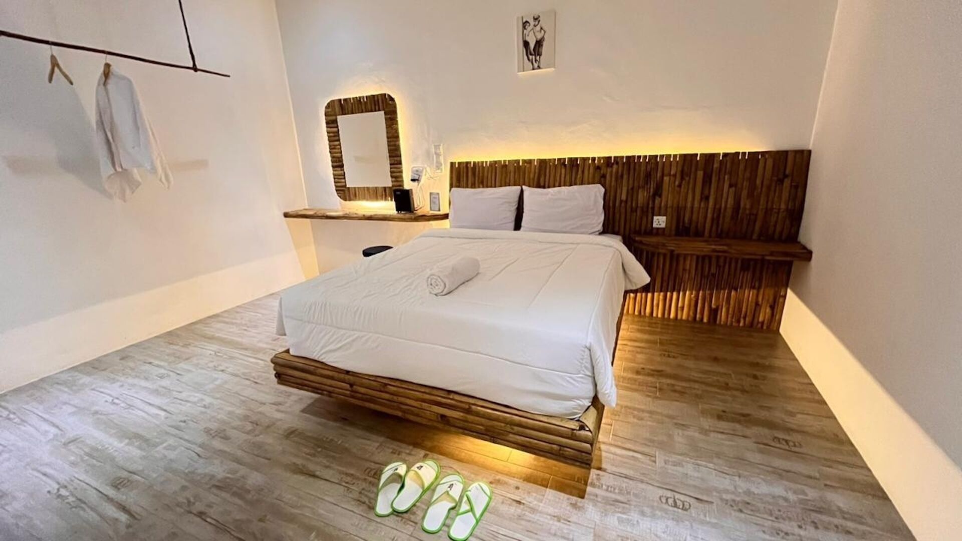 A bedroom with a bed, wooden headboard, bedside table, wall-mounted mirror, and a pair of slippers on the floor.