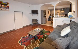 A living room with a sofa, a patterned rug, a television, and a kitchen area visible through an archway. - Appart. dans maison de village (Montagnac-Montpezat)