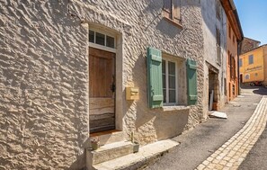 A narrow street with a building featuring a wooden door and green shutters, a small step leading to the entrance, and a paved sidewalk. - Appart. dans maison de village (Montagnac-Montpezat)