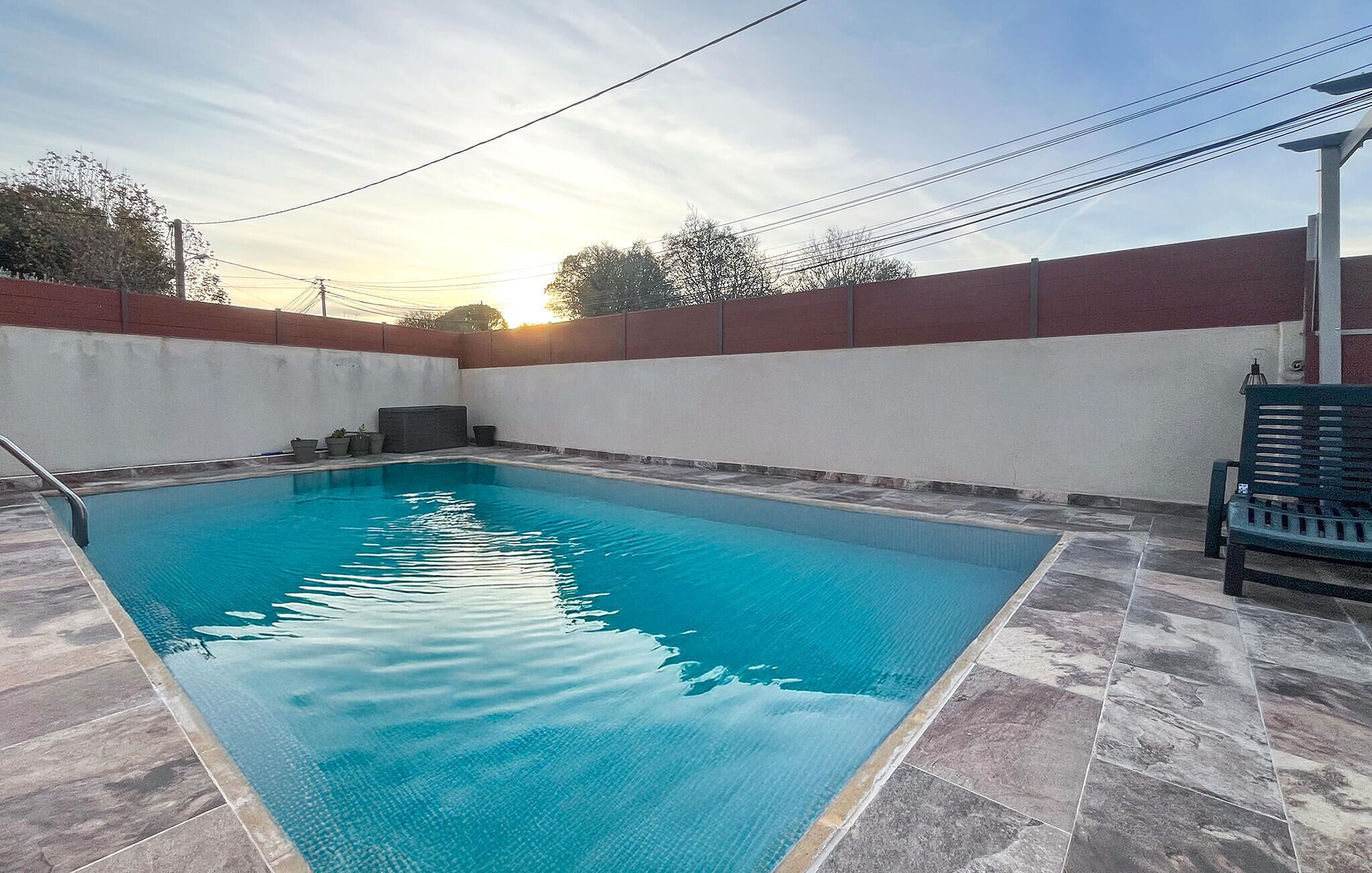 Una piscina rectangular con plataforma azulejada, rodeada por un muro de ladrillo bajo y una valla blanca.