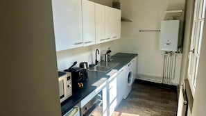 A compact kitchen with white cabinets, a built-in washing machine, and a water heater. - La Propriété de Bacchus - Écrin de Loire - Design & Confort (Saumur)