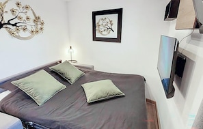Ein Schlafzimmer mit Bett, Kissen, einem an der Wand montierten Fernseher und einem gerahmten Kunstwerk.