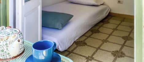 Una habitación pequeña con una cama individual, una ventana, una mesa con una tetera y una taza, y un suelo de baldosas.