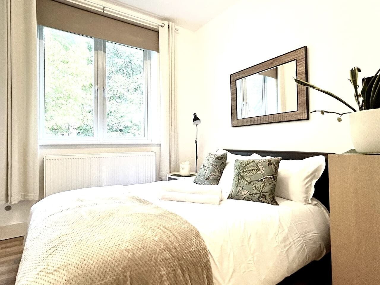Une chambre avec une grande fenêtre, un lit recouvert de linge de lit blanc, une table de chevet avec une lampe et un miroir accroché au mur.