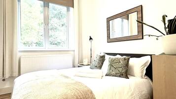 Une chambre avec une grande fenêtre, un lit recouvert de linge de lit blanc, une table de chevet avec une lampe et un miroir accroché au mur.