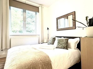 A bedroom with a large window, a bed with white bedding, a bedside table with a lamp, and a mirror on the wall. - Chic Kensington Olympia home, 2bed/2bath for a perfect London stay! (Greater London)