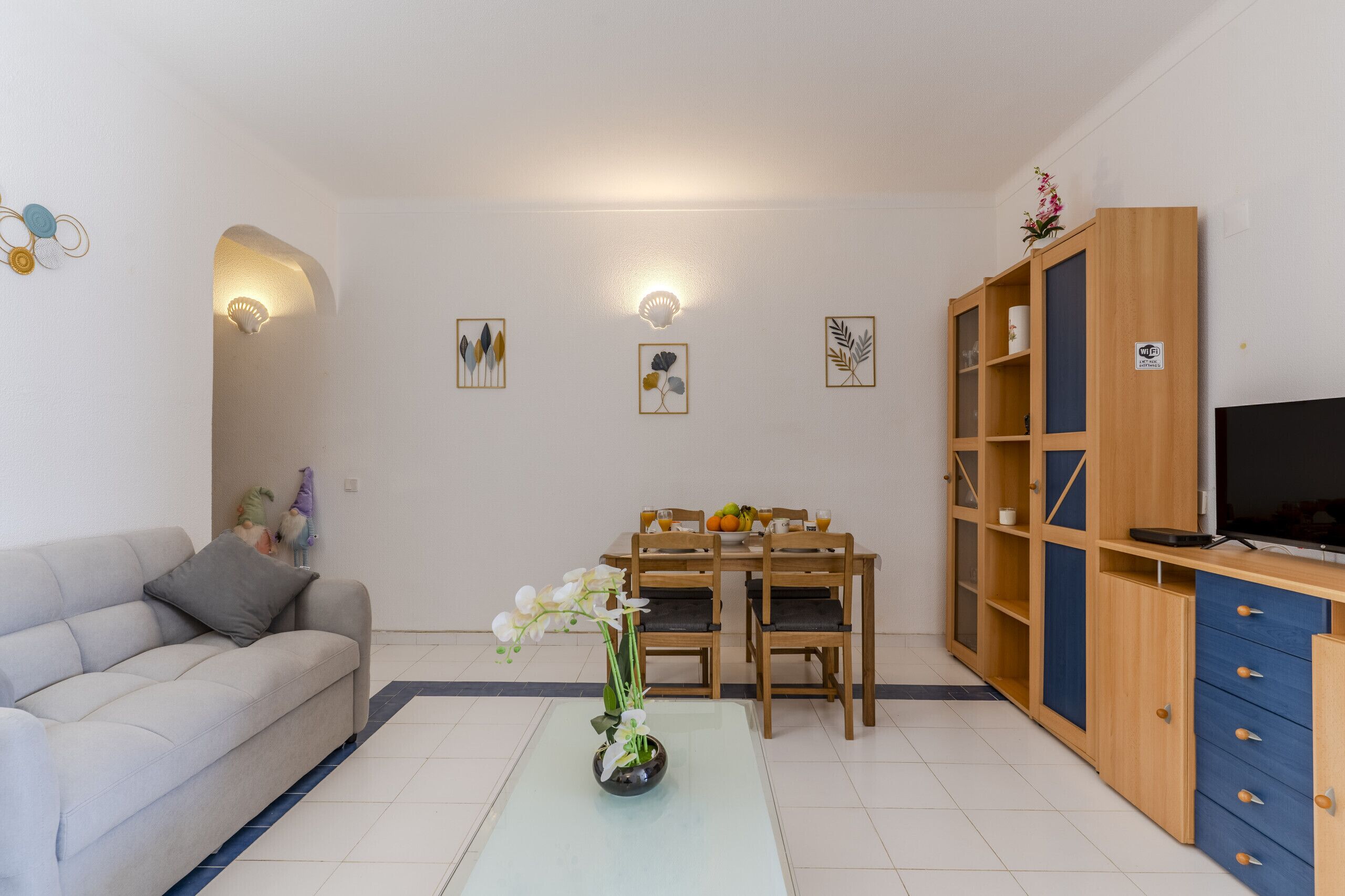 A modern living room with a sofa, a dining table with chairs, a coffee table with flowers, a TV stand with a TV, and a bookshelf.