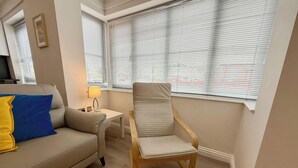 Una sala de estar con sofá, una silla, una mesa con lámpara y ventanas amplias con persianas.