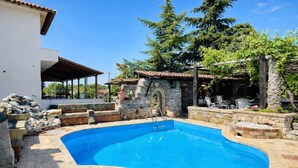A swimming pool with a stone wall and a covered patio area. - Saltos Village Apartment - Serene Poolside Escape (Nea Propontida)