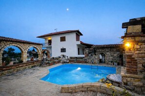 A pool area with a stone archway, a white building with a red roof, and a stone wall with a light fixture. - Saltos Village Apartment - Serene Poolside Escape (Nea Propontida)
