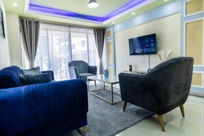 A modern living room with a blue sofa, a black armchair, a coffee table, and a flat-screen TV mounted on the wall. - Crestpark View Palace Apartment (Nairobi)