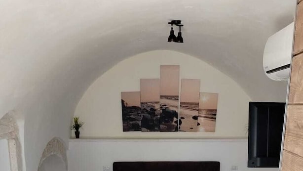 A bedroom with a stone floor, a bed with a headboard, a wall-mounted TV, and a small table with a lamp.