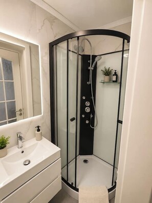 A modern bathroom with a glass-enclosed shower, a white sink with a mirror, and a small shelf with toiletries. - Luxury Designer Studio | King Bed | Near Old Town (Bucharest)