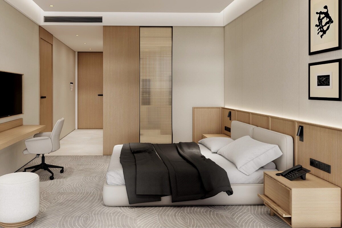 A modern hotel room with a bed, bedside table, desk, chair, and a large mirror.