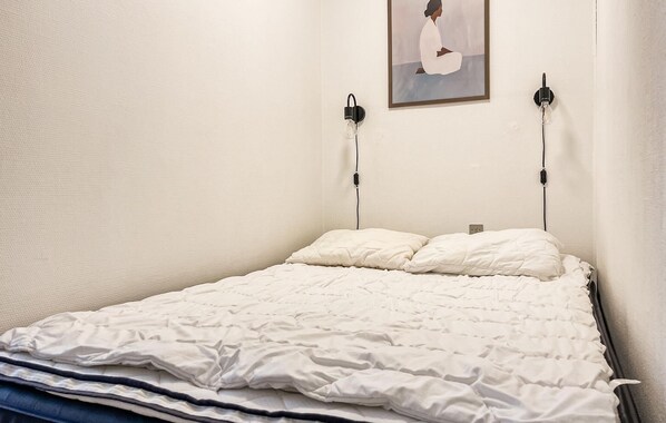 Uma cama bem arrumada com um edredom branco e almofadas, uma luminária fixada na parede e um quadro emoldurado.