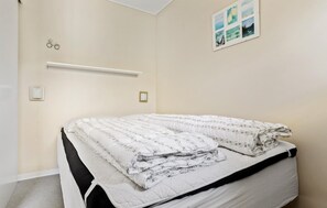 Ein ordentlich bezogenes Bett mit grau-weiß gestreifter Bettdecke und Kissen, ein Nachttisch mit Lampe sowie ein an der Wand hängendes Bildergestell mit mehreren Bildern.