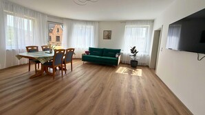A spacious living room with a dining area, wooden flooring, a green sofa, and a flat-screen TV mounted on the wall. - Zum Rheinberg – Balcony Suite (Braubach)
