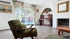 A living room with a green armchair, a piano, a fireplace, and a large window with curtains. - Charming Villa for 6 in Agde (Agde)