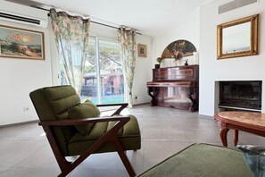 Ein Wohnzimmer mit einem grünen Sessel, einem Klavier, einem Kamin und einem großen Fenster mit Vorhängen.