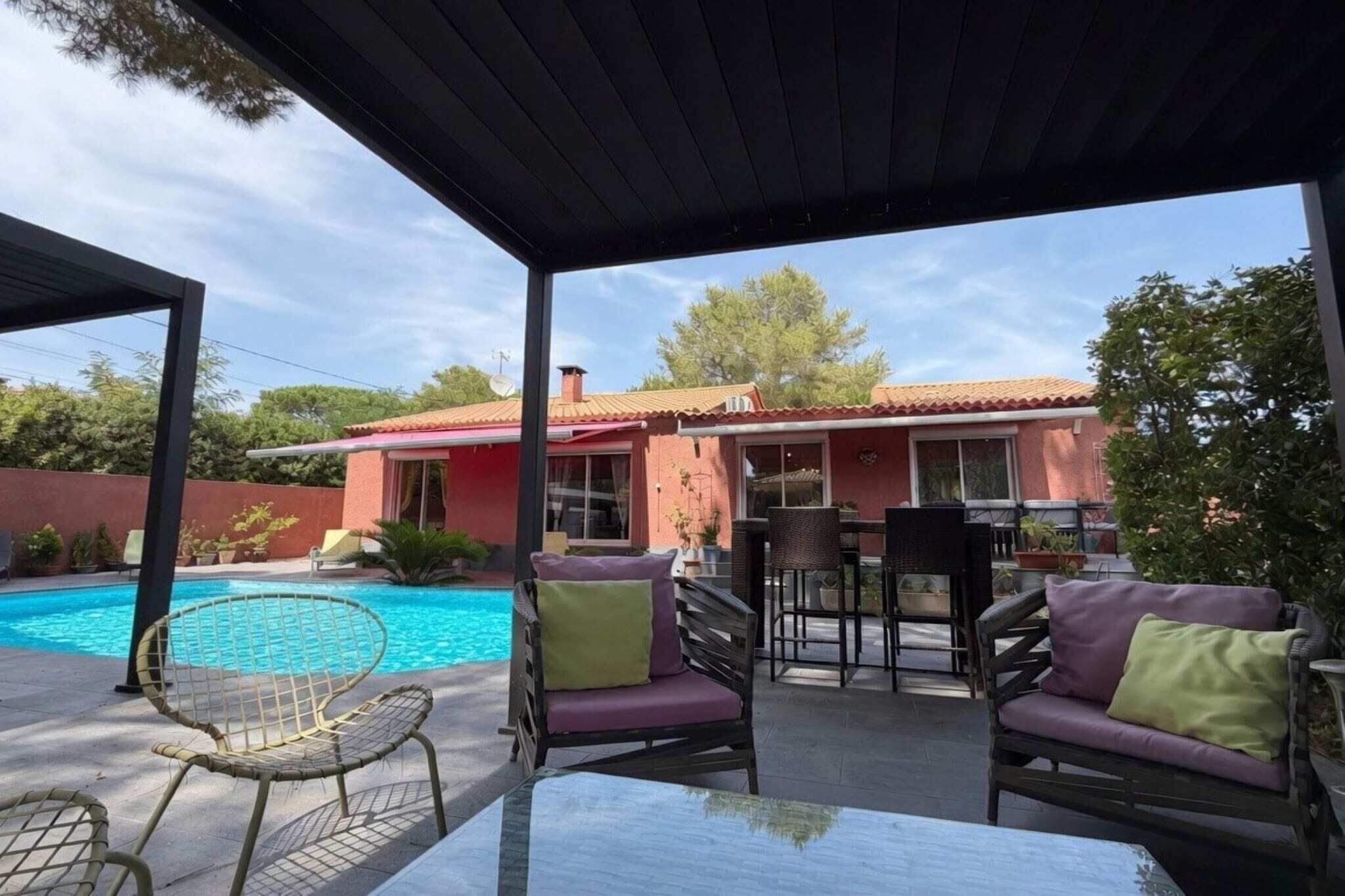 Un área junto a la piscina con un porche cubierto, mobiliario exterior y una casa al fondo.