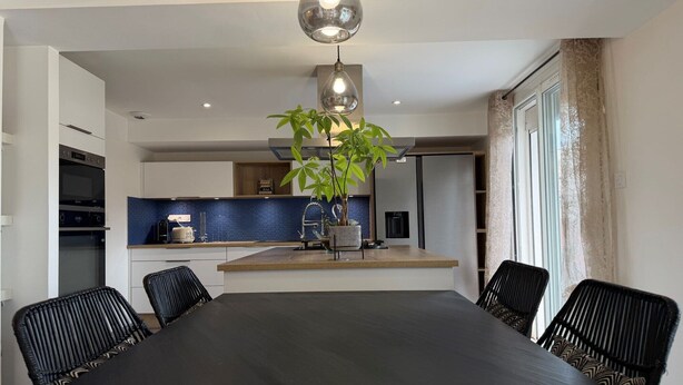A modern dining area with a long table, black chairs, a hanging light fixture, and a kitchen in the background. - Charming Villa for 6 in Agde (Agde)