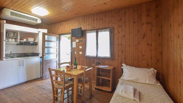 Habitación pequeña con detalles de madera, una kitchenette, una mesa de comedor con sillas, una cama con almohadas y una ventana con cortinas.
