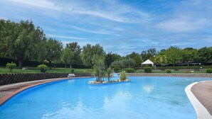 Una piscina con un paseo de ladrillo alrededor, árboles y un pabellón blanco al fondo.