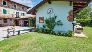 Um edifício rústico com telhado de telhas, um pequeno jardim com um banco e uma parede branca com um ícone religioso.
