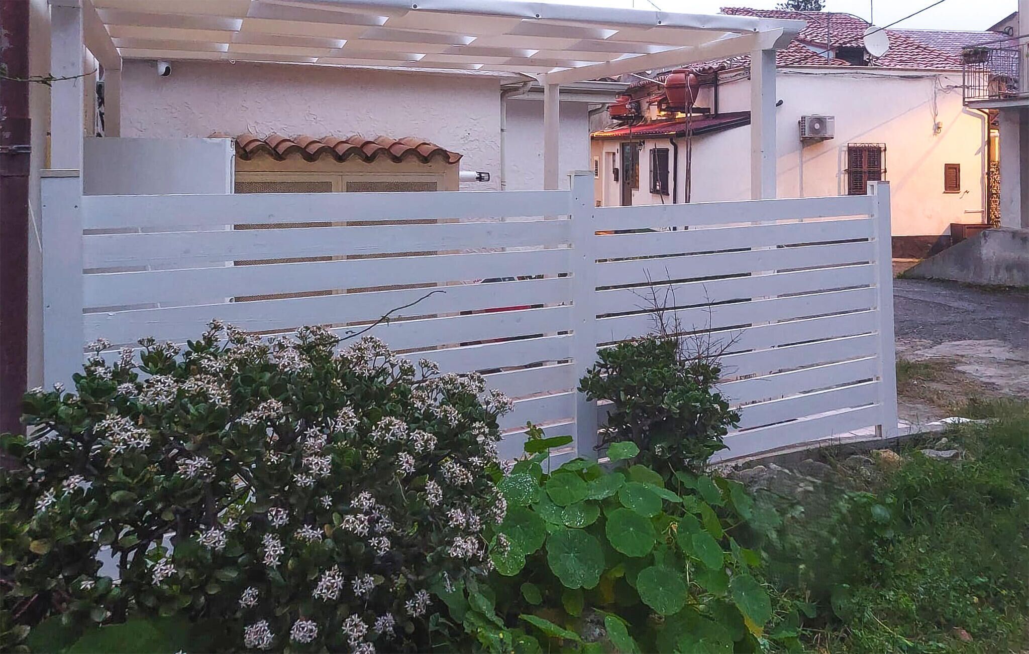 Una reja metálica blanca con listones horizontales, una pared blanca con tejado de baldosas y un arbusto pequeño con flores blancas al frente.