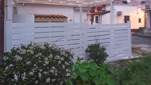 A white metal gate with horizontal slats, a white wall with a tiled roof, and a small bush with white flowers in the foreground. - Lovely apartment in Tropea with WiFi (Tropea)