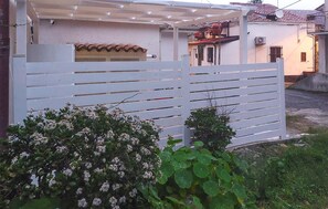 Ein weißes Metallgitter mit horizontalen Lamellen, eine weiße Mauer mit einem Ziegeldach und ein kleiner Strauch mit weißen Blüten im Vordergrund.