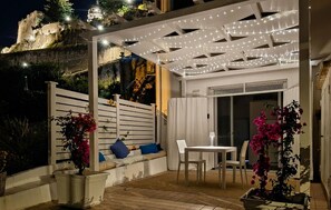 Eine Terrasse mit Tisch und Stühlen, eine überdachte Sitzecke mit Kissen und ein Blick auf ein historisches Gebäude im Hintergrund.