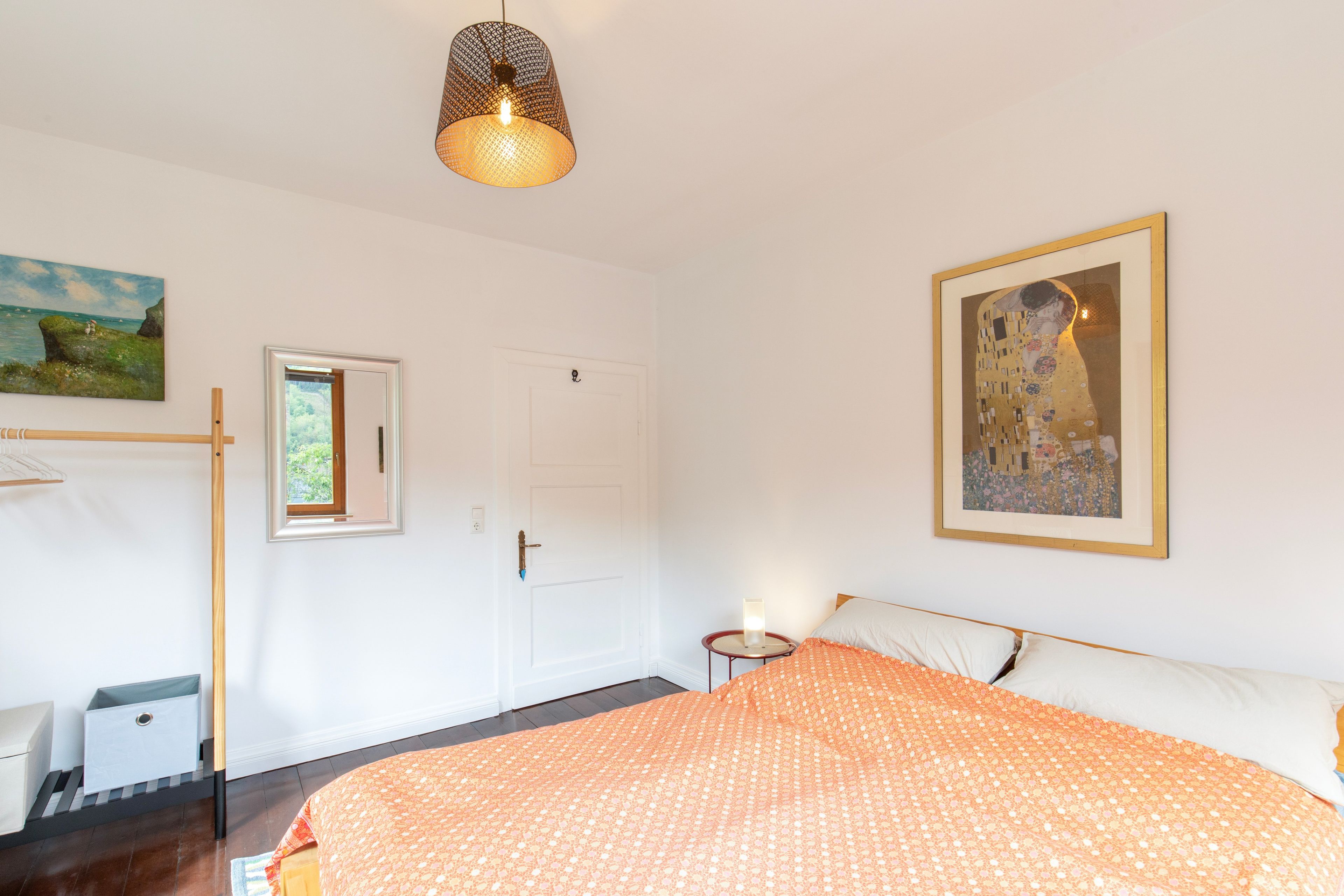 A bedroom with a bed, a door, a window, a painting, and a hanging light fixture.
