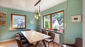 A dining area with a wooden table and chairs, large windows with views of greenery, and wall-mounted light fixtures. - Holiday Home 'Hiwwelhaus' with Private Terrace, Private Garden and Wi-Fi (Oberdiebach)