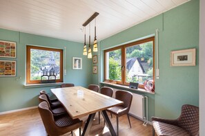 A dining area with a wooden table and chairs, large windows with views of greenery, and wall-mounted light fixtures.