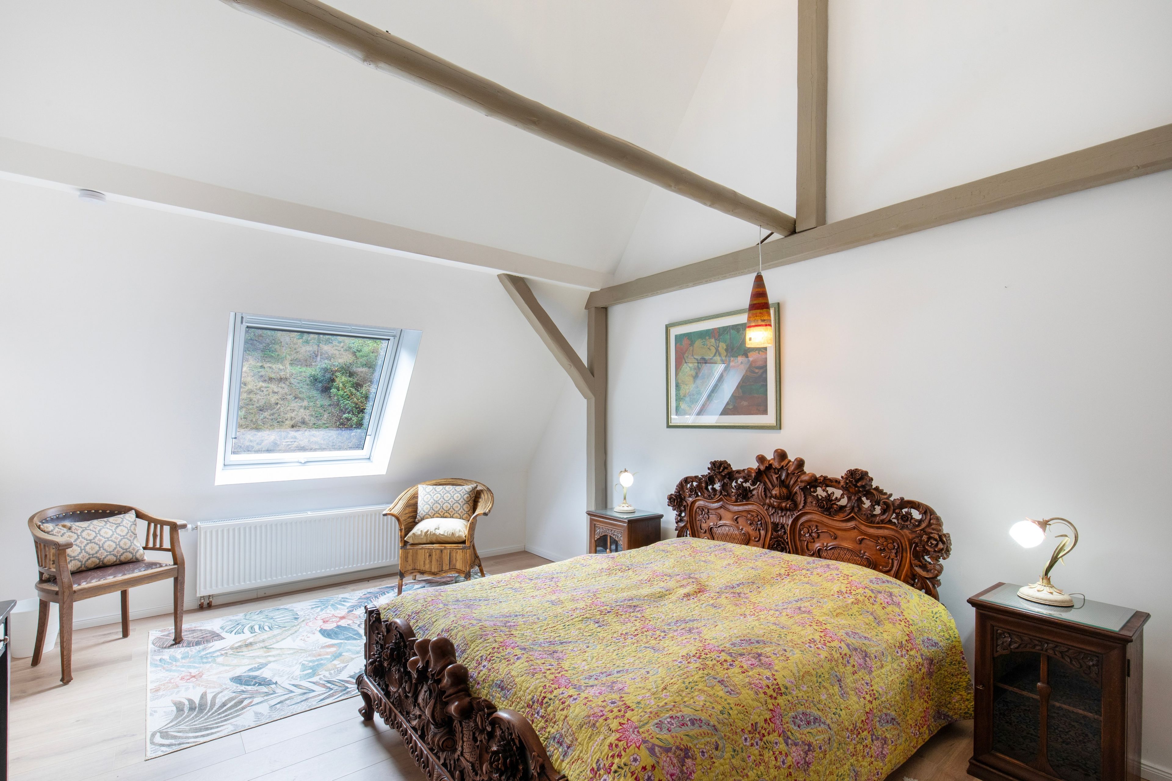 Ein Schlafzimmer mit einem großen Bett, einem Fenster, einem Sessel, einem kleinen Tisch und einer Lampe.
