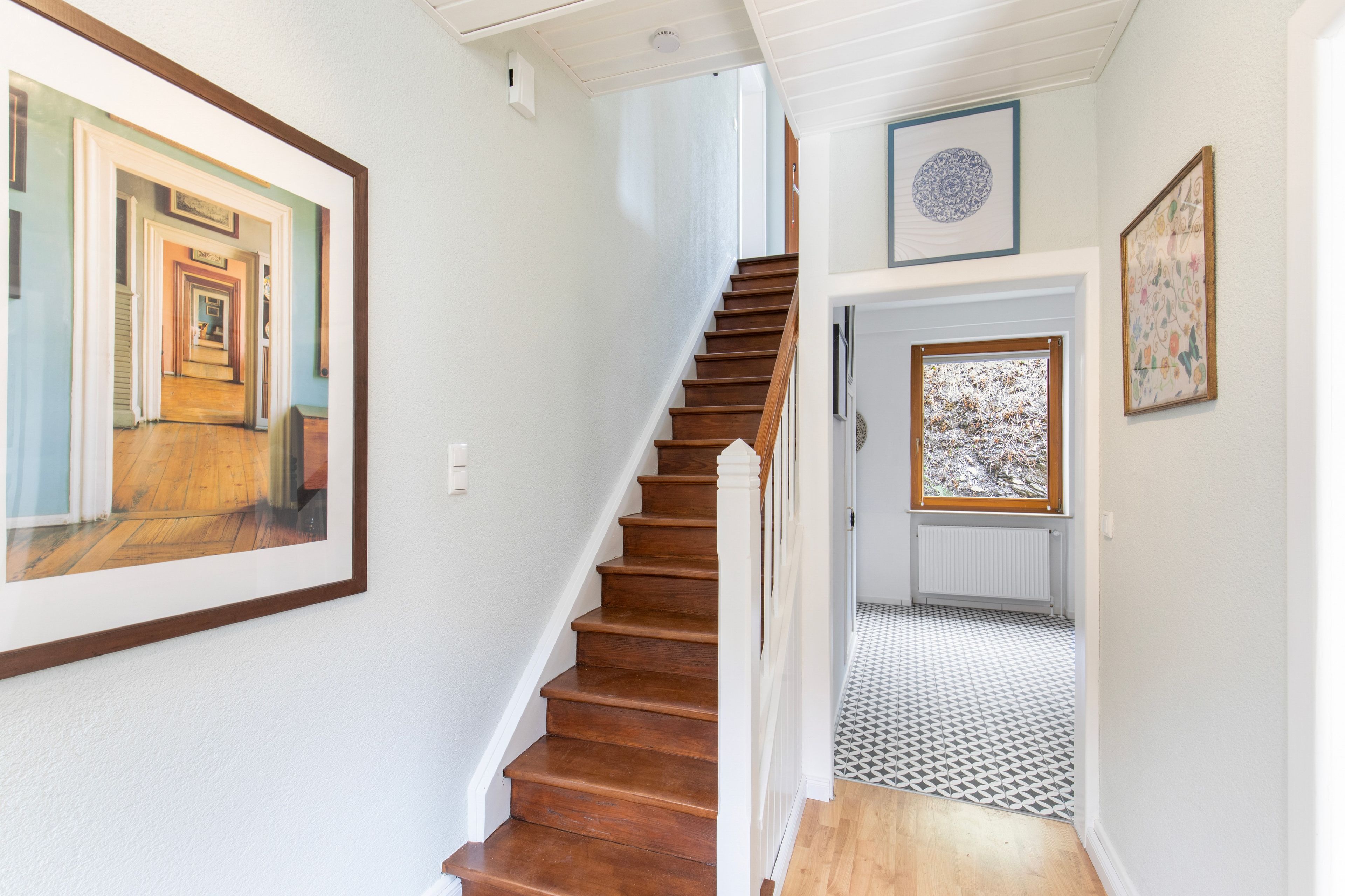 Un escalier avec des marches en bois menant à un palier, un mur blanc orné d’un tableau encadré et une porte avec une fenêtre.