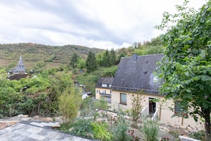 A residential area with a house featuring a sloped roof, a garden with various plants, and a hill with trees in the background.