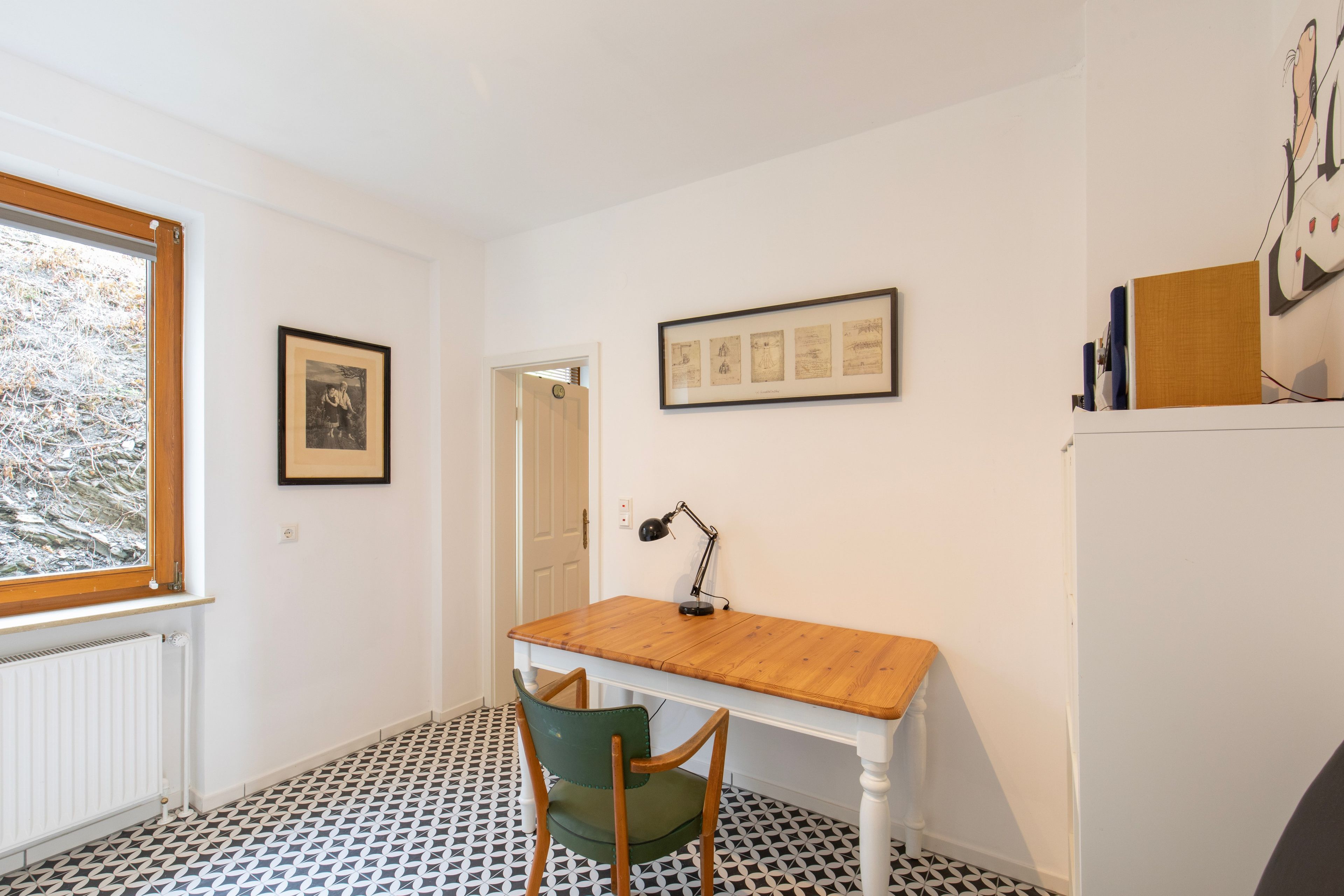 Een kamer met een houten bureau, een groene stoel, een radiator, een raam met uitzicht op bomen en een muur met ingelijste foto's.