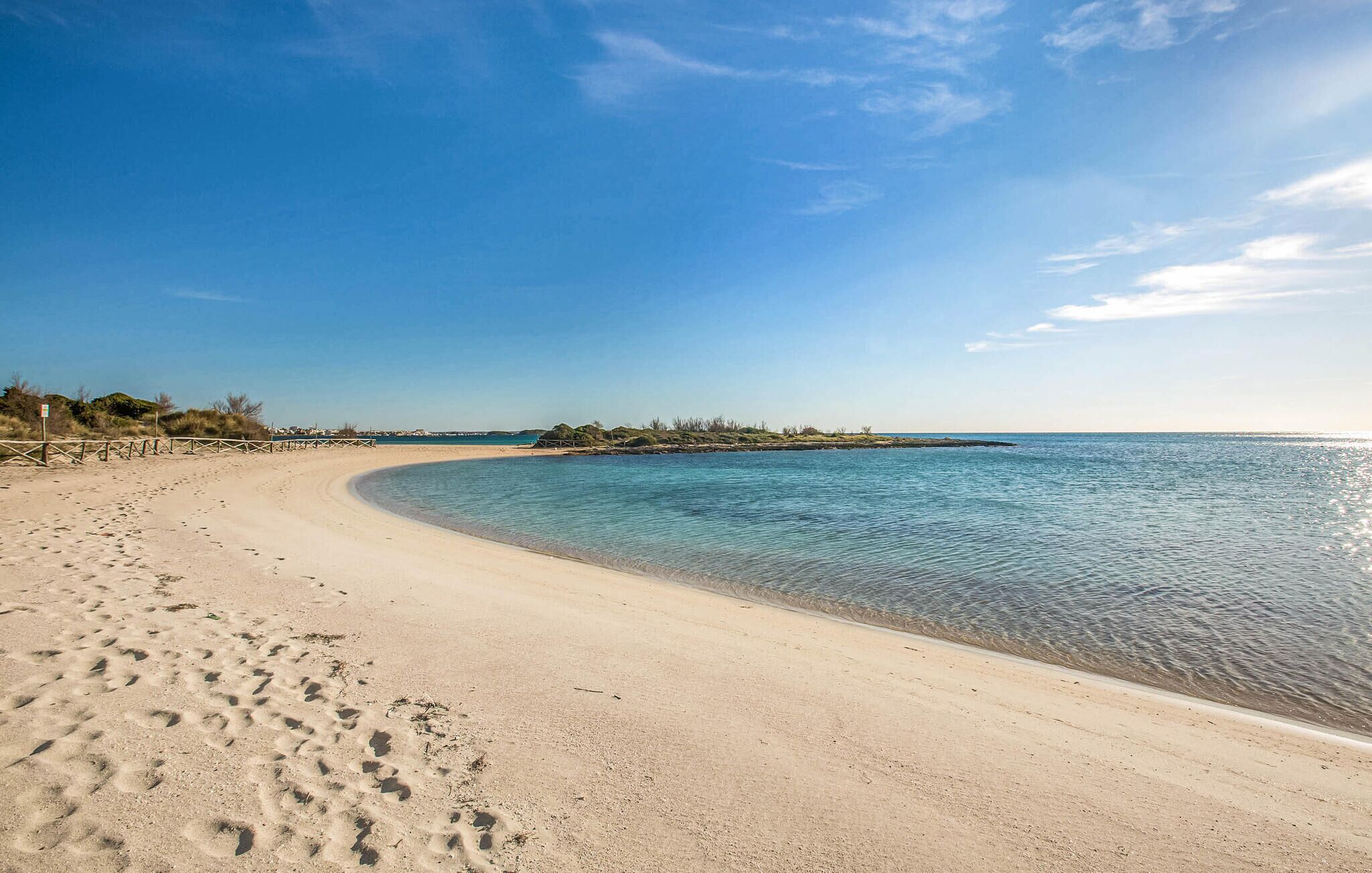 Uma praia arenosa com pegadas que se estendem até a água, céu claro e mar calmo.