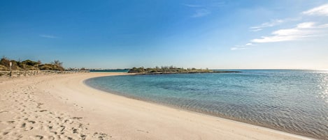 En sandstrand med fodaftryk, der fører ned til vandet, en klar himmel og et roligt hav.