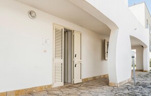 Ein Gebäude mit heller Fassade, zwei Holztüren mit Fensterläden, einem gepflasterten Eingangsbereich und blauem Himmel.