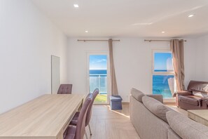A modern living room with a wooden dining table, a sofa, and a chair. There are two windows with curtains, and a view of the sea is visible through the windows. - 2BR La Furnia Home - Balcony & Natural Pool (Gáldar)