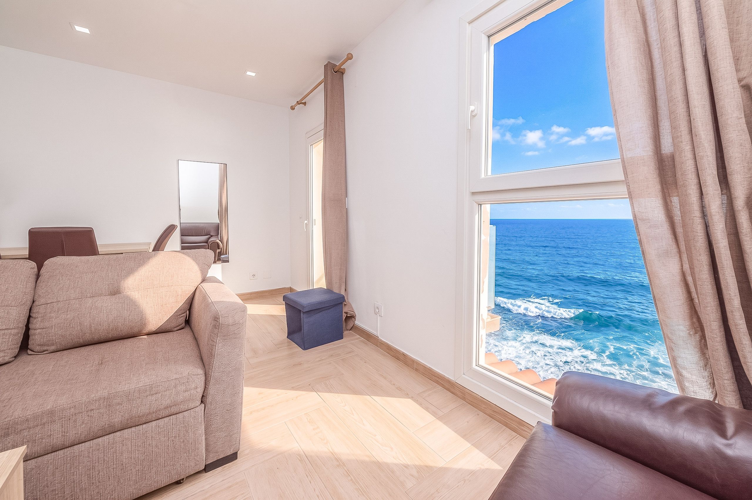 A modern living room with a large window offering a view of the sea, a sofa, a small table, and a mirror.